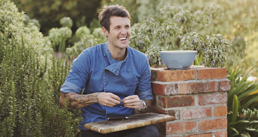 Ben Milbourne sitting outdoors preparing fresh ingredients with a chopping board on his lap